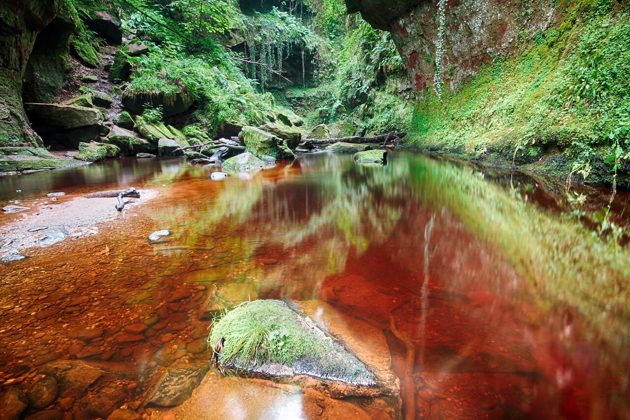 Blood-red water pools between green moss-covered sandstone walls in Finnich Glen, a mossy boulder rising from the shallows.