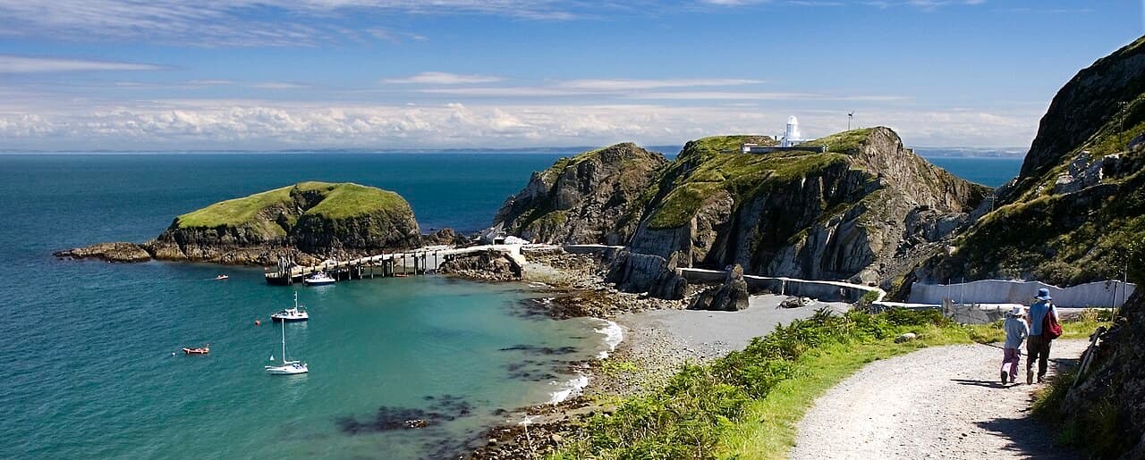 Yachts moored in the turquoise Landing Bay of Lundy, with the island track climbing past the jetty towards the white lighthouse on the headland.
