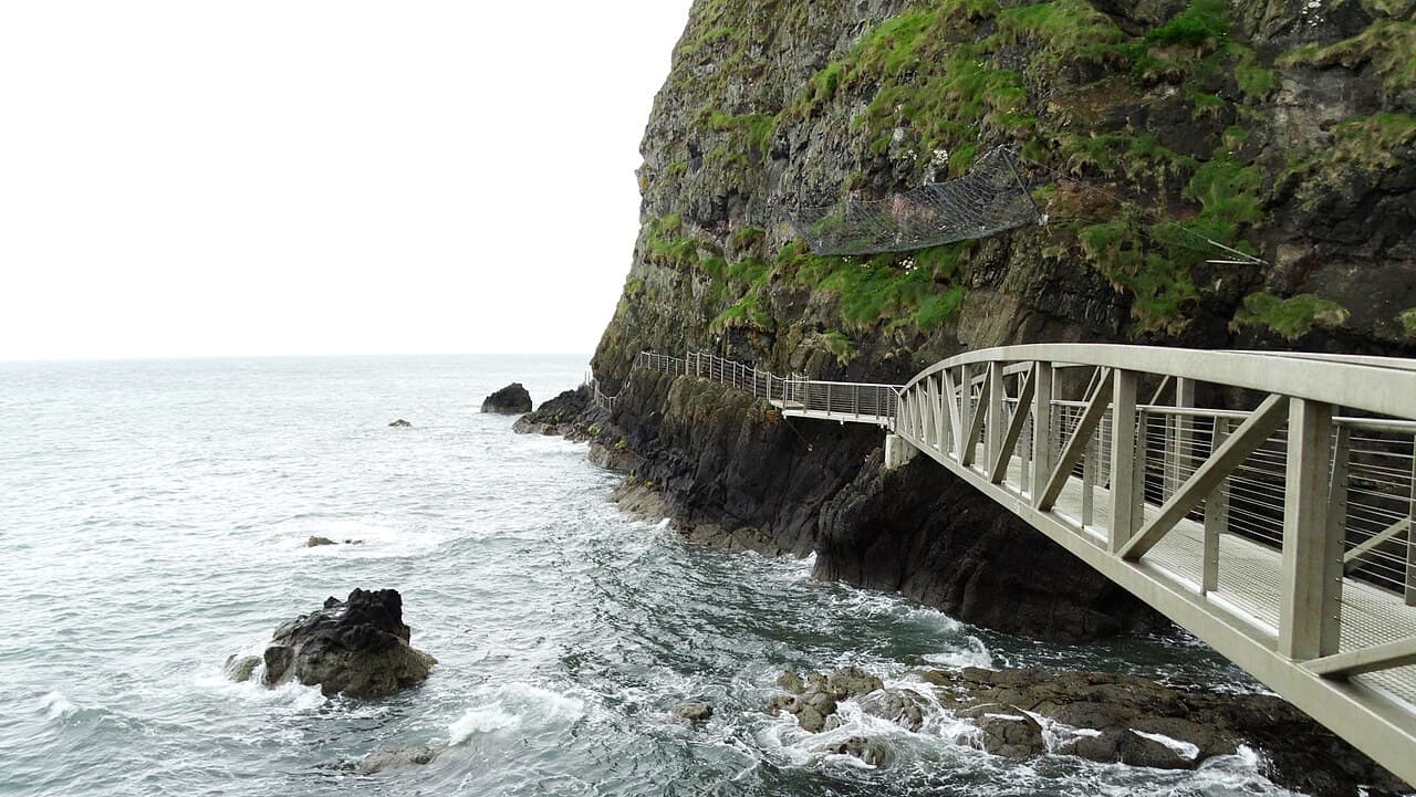 A pale iron bridge of the Gobbins cliff path curves around the foot of a black basalt cliff above the surging sea.
