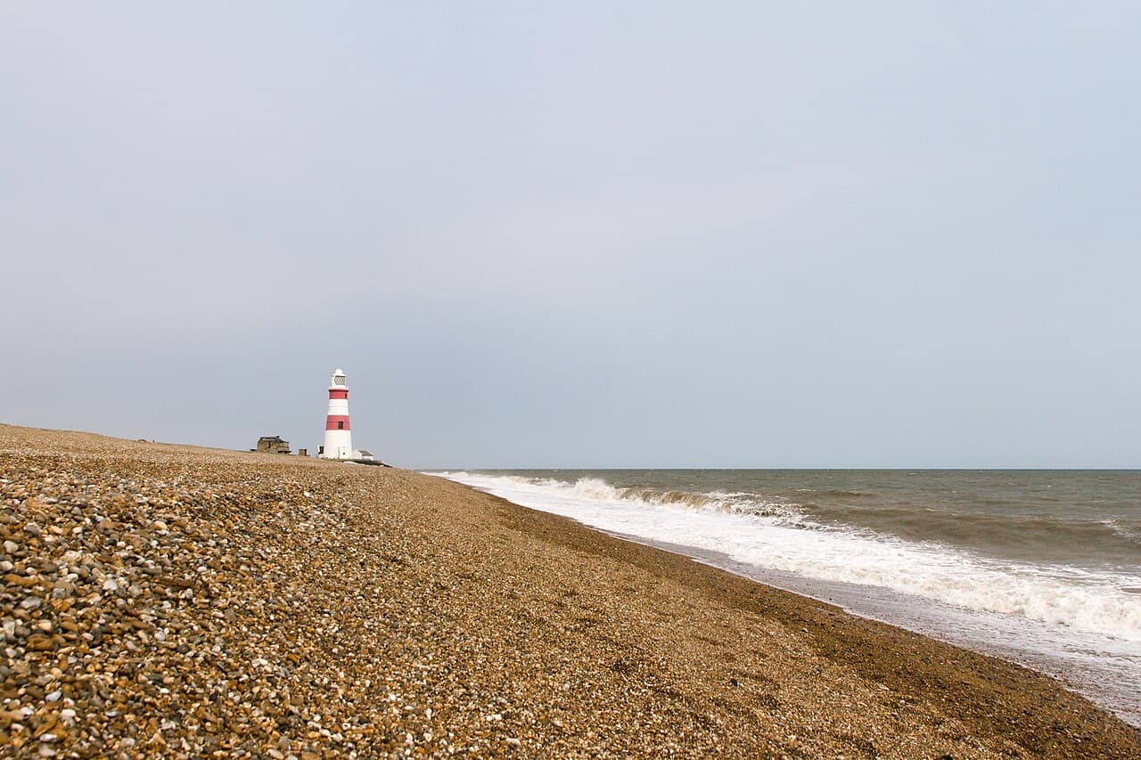 The red and white Orfordness lighthouse stands alone on the shingle beach as waves break along the edge of the spit.