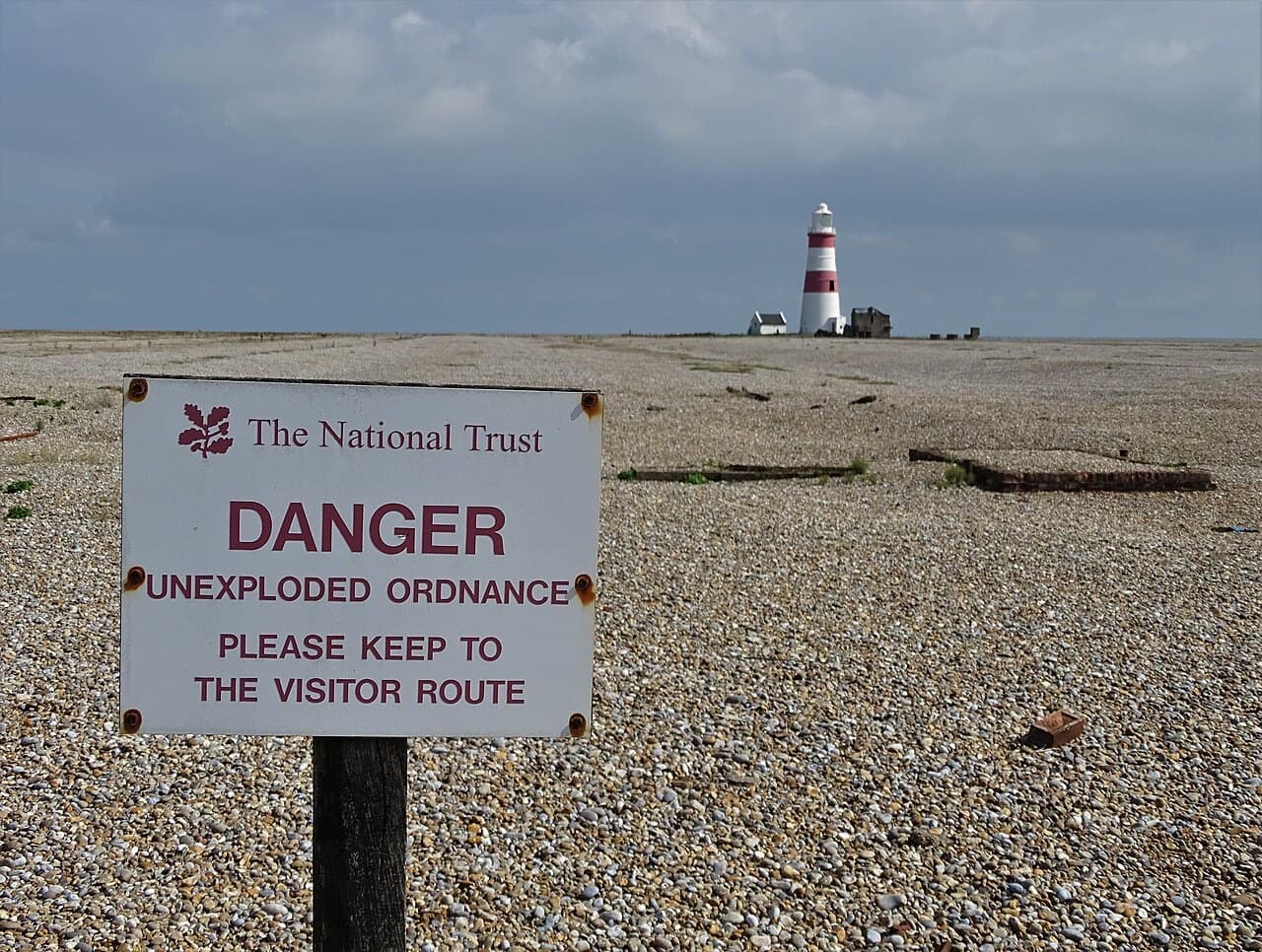 A National Trust sign on the open shingle of Orford Ness warns of unexploded ordnance, with the lighthouse on the horizon behind.