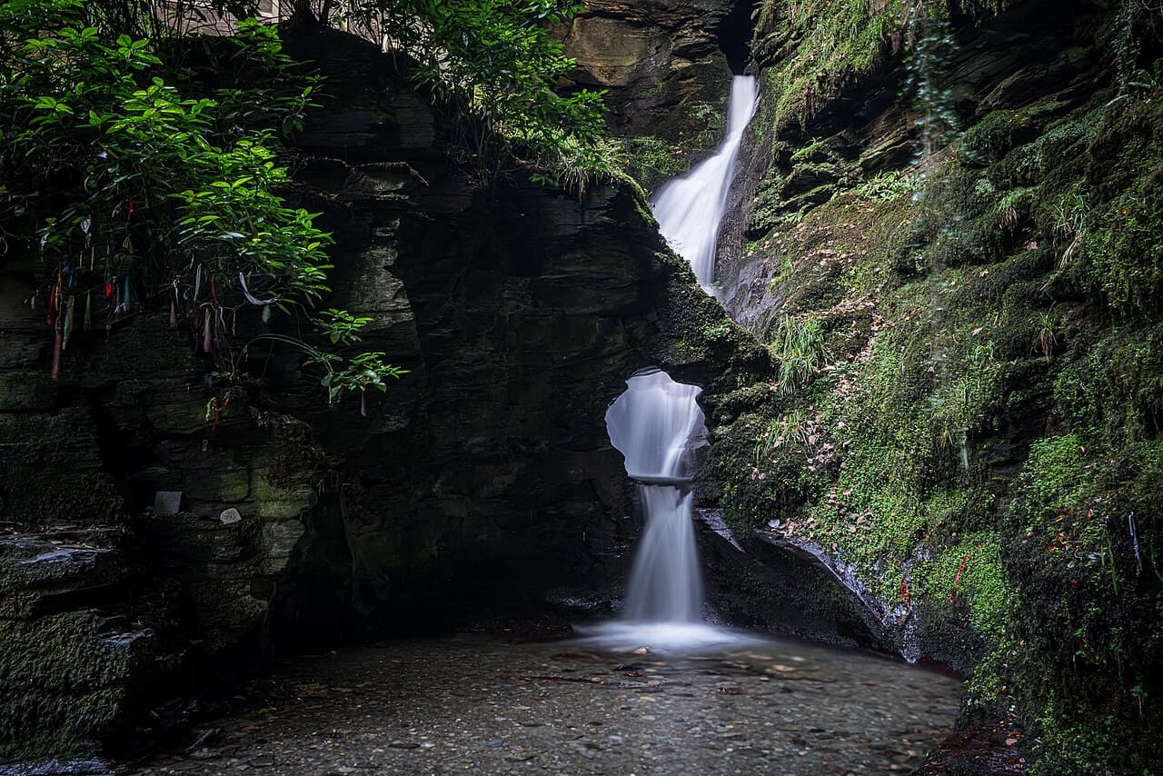 The full double cascade of St Nectan's Kieve seen across its dark basin, with prayer ribbons tied to a tree on the mossy left-hand cliff.