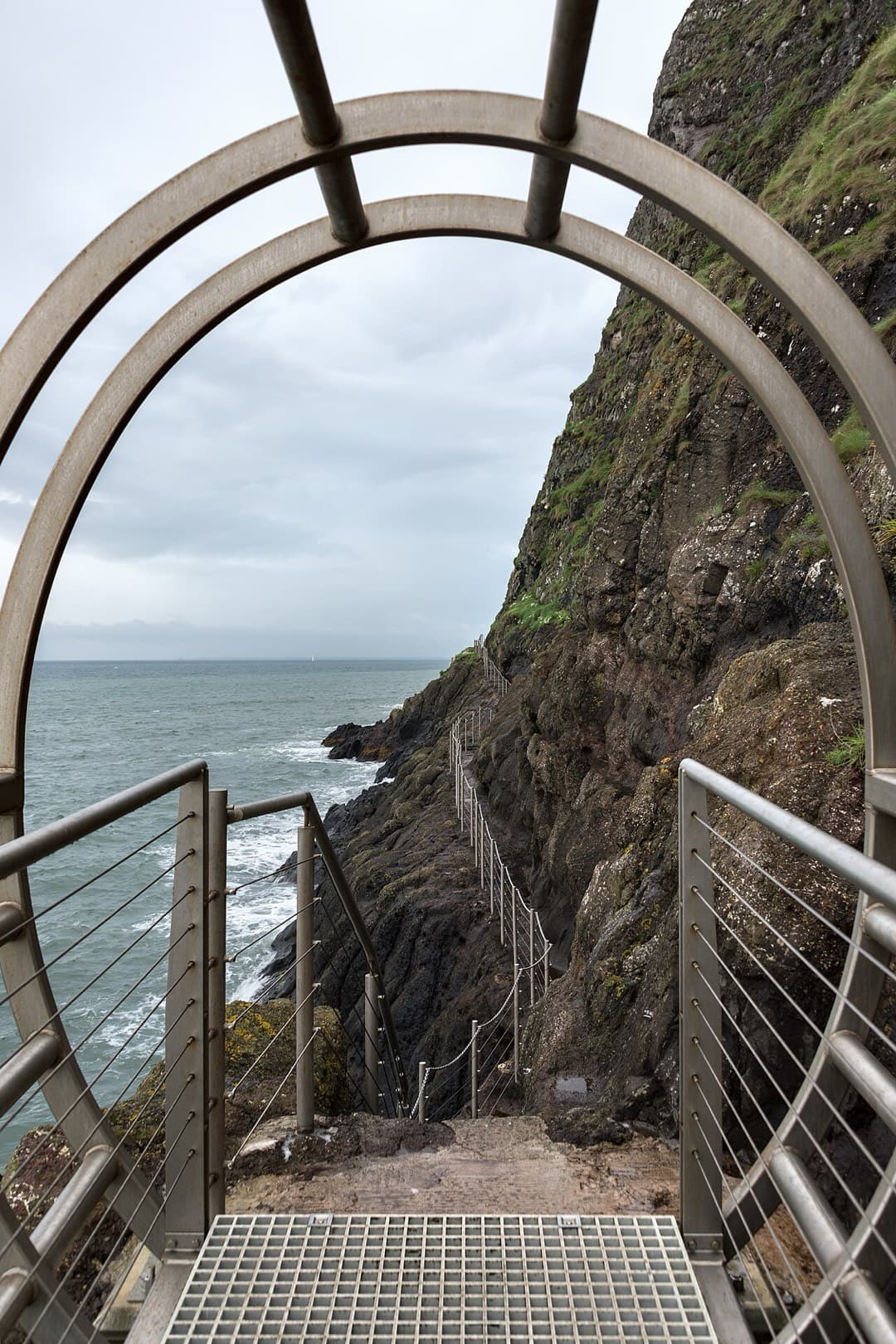 Seen through the round mouth of a tubular bridge, railed steps of the Gobbins path drop steeply along the cliff toward the waves.