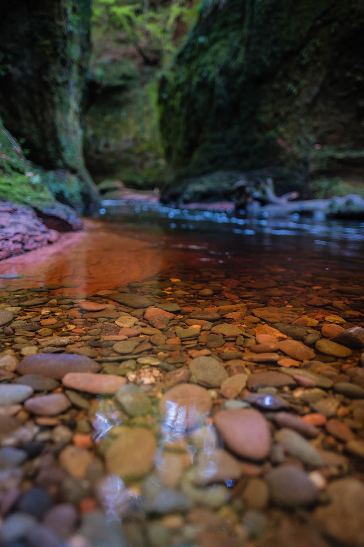 Red and brown pebbles beneath the clear peat-stained water of the Carnock Burn, the mossy walls of Finnich Glen blurred beyond.