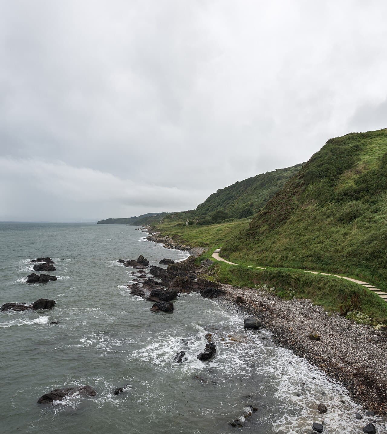 The approach path to the Gobbins winds along a stony shore beneath green slopes on the Islandmagee coast under a grey sky.