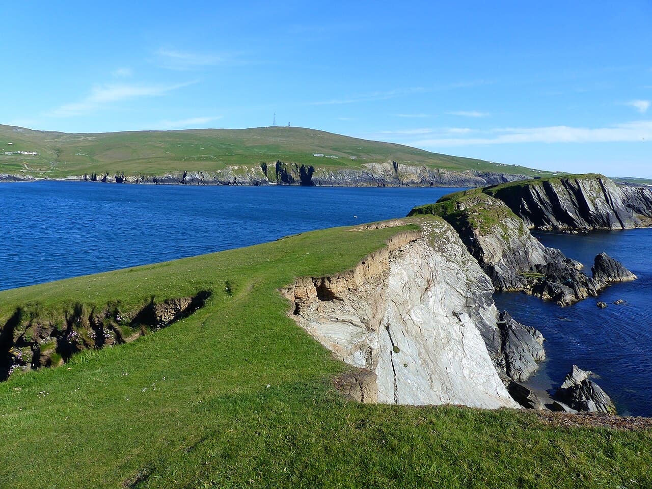 Green clifftops fall away to stacks and headlands of dark rock in a deep blue sea on the seaward side of St Ninian's Isle.