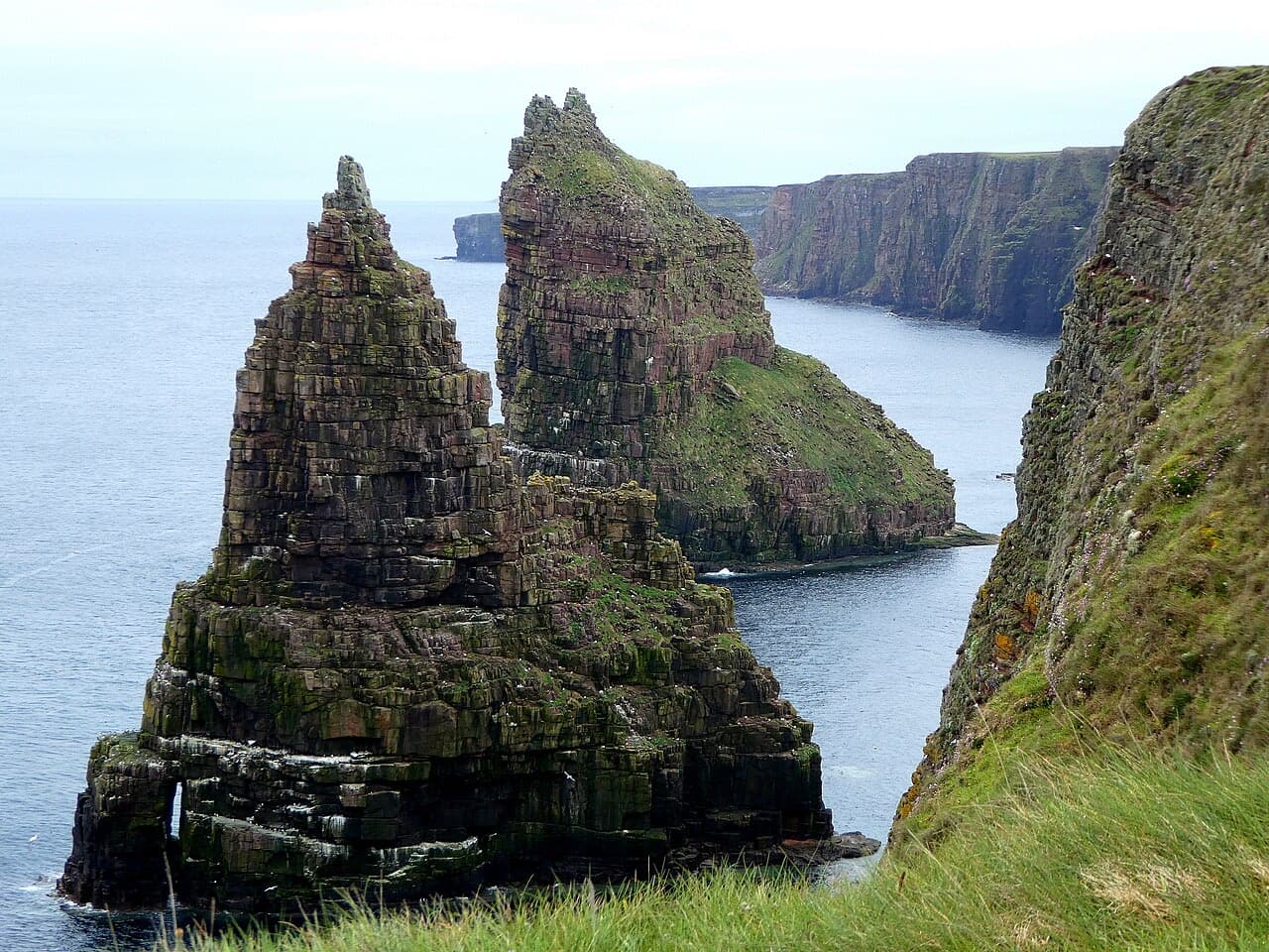The twin jagged Stacks of Duncansby rise from the sea beside banded red sandstone cliffs on the Caithness coast.