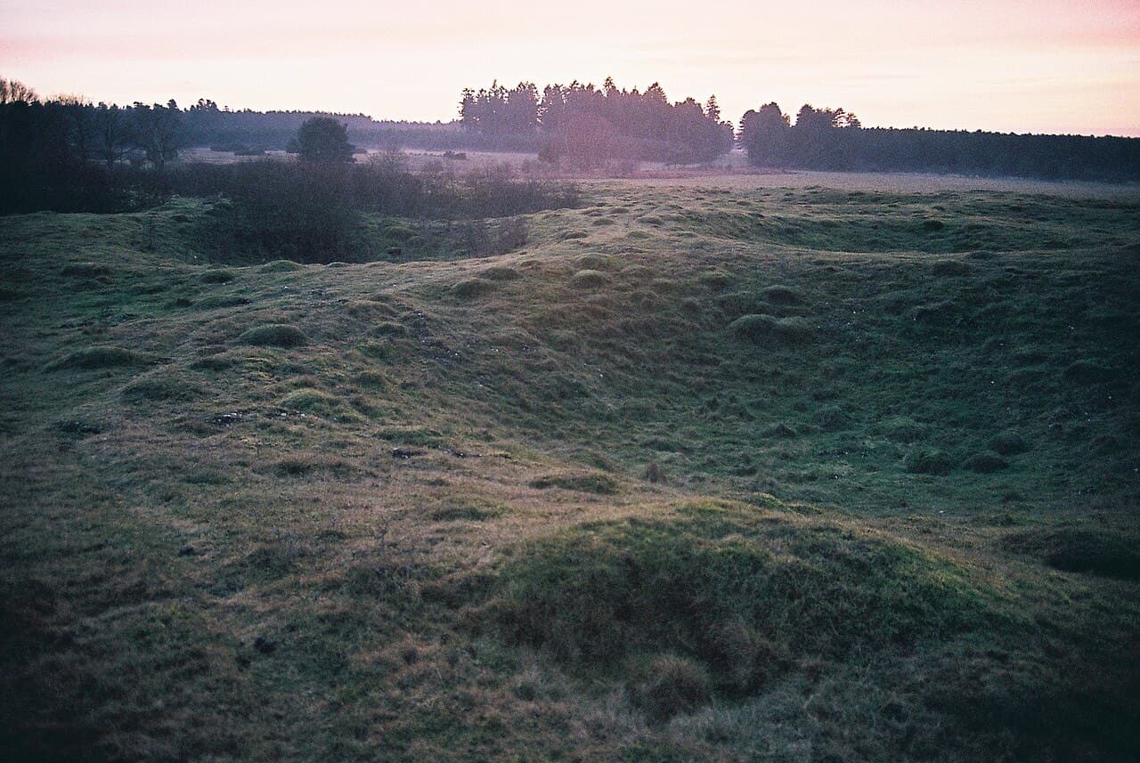 The pitted grassland of Grime's Graves at dusk, hollows of filled-in flint mineshafts dimpling the field under a pink sky.