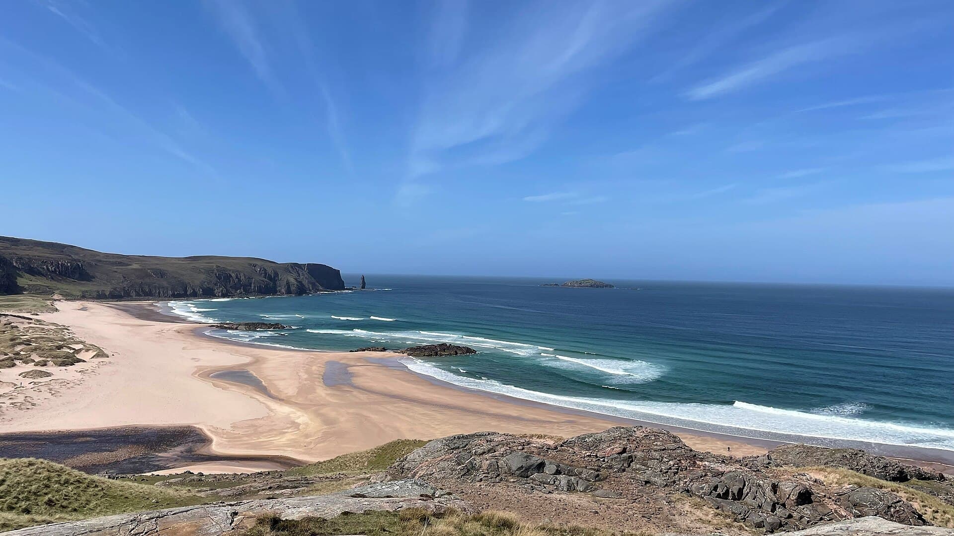 Elevated view over the mile-long curve of Sandwood Bay, pink-tinged sand and turquoise surf sweeping toward dark cliffs and the distant sea stack Am Buachaille.
