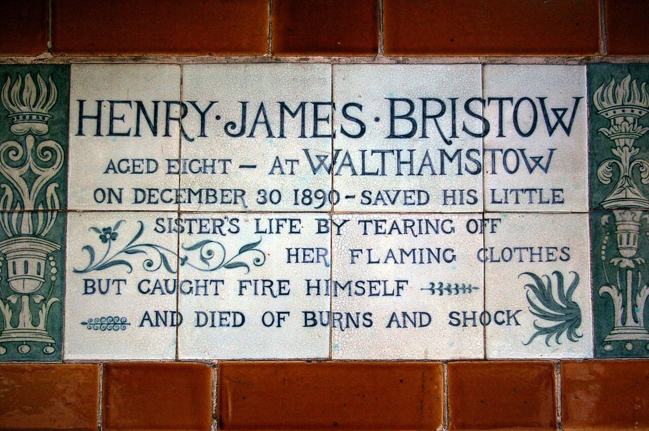 Hand-lettered Victorian ceramic tablet framed by terracotta tiles, commemorating eight-year-old Henry James Bristow, who saved his little sister's life by tearing off her flaming clothes.