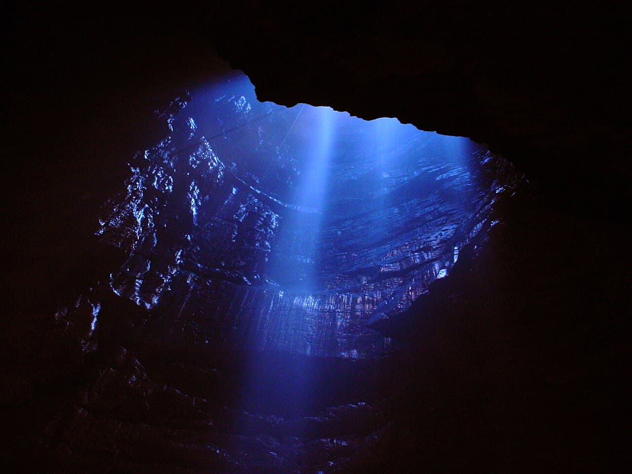 A pale blue shaft of daylight pours down the flooded surface shaft of Gaping Gill, silvering the wet limestone walls of the black main chamber.
