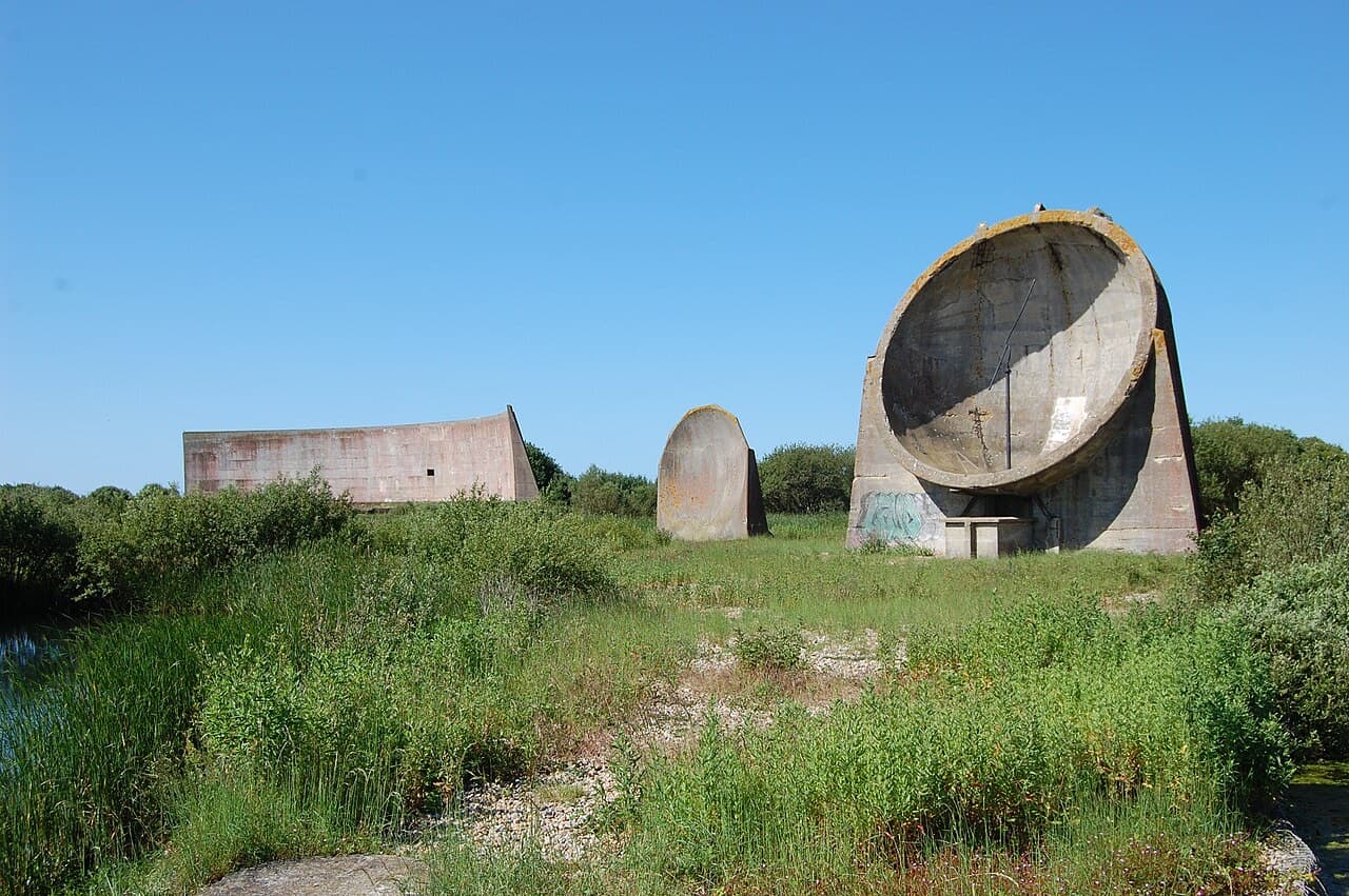 All three concrete sound mirrors at Denge, the long wall, the small dish and the great bowl, stand in rough scrub under a clear sky.