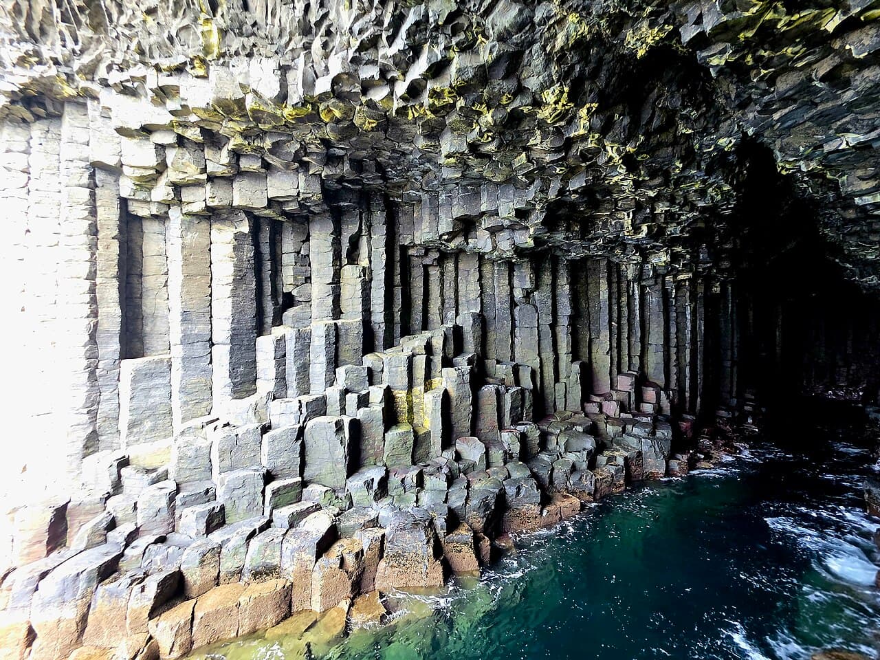 Hexagonal basalt columns step down into clear emerald seawater inside Fingal's Cave, beneath a ceiling flecked with gold-green lichen.
