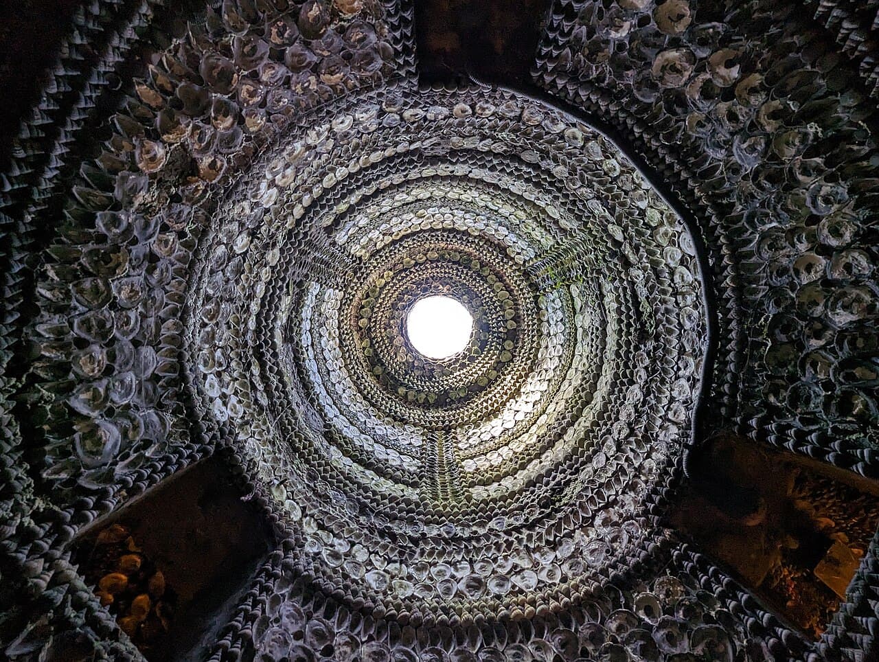 View straight up into the grotto dome, where concentric rings of shells spiral around a small skylight pouring in daylight.