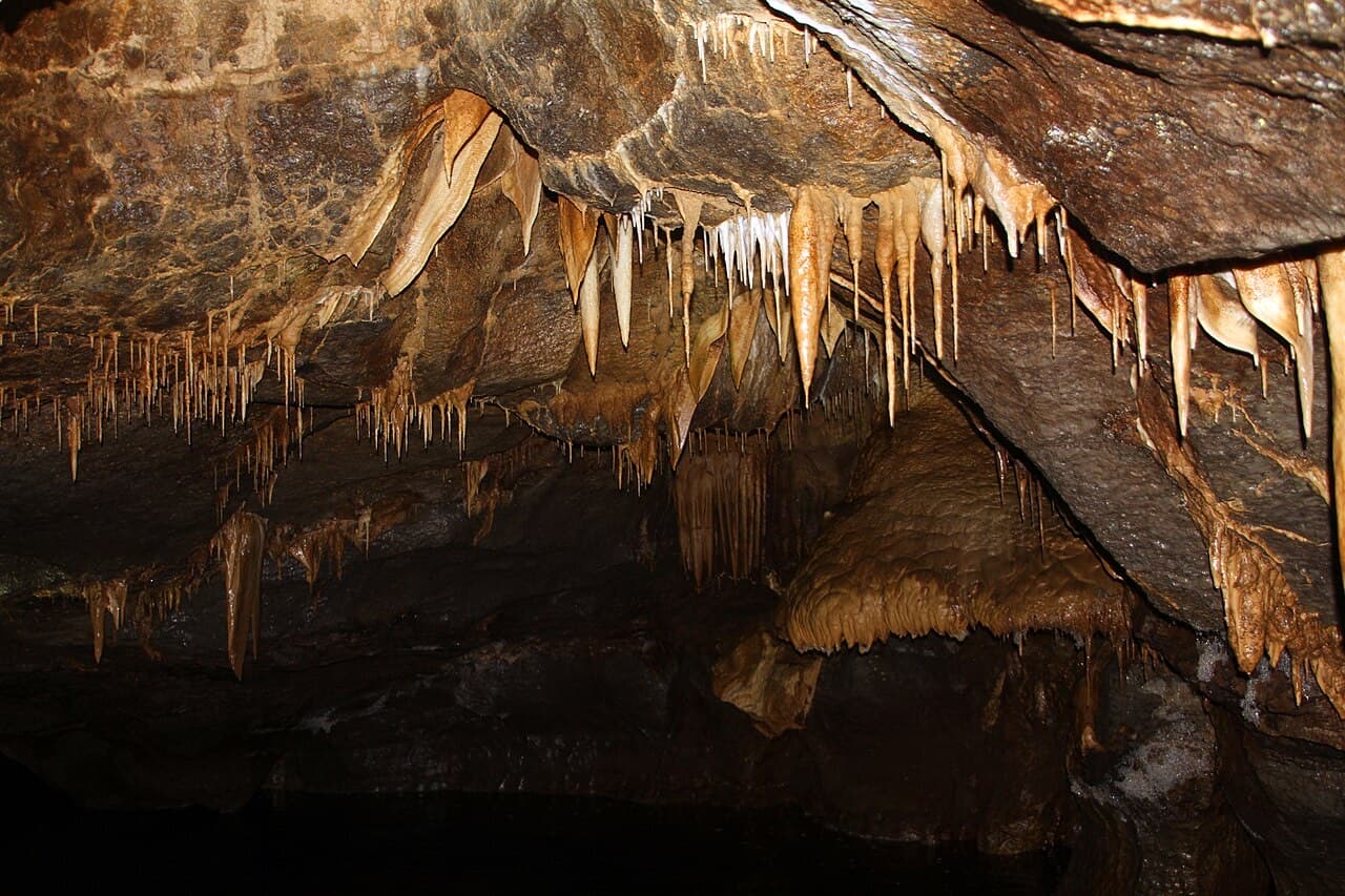 Stalactites fringe the roof of a broad chamber in the Marble Arch Caves, hanging above dark water on the cavern floor.