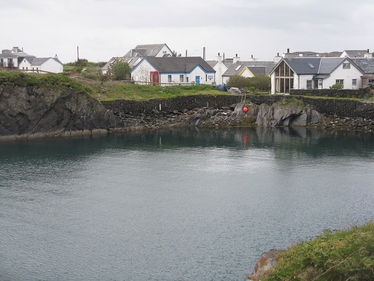 A row of whitewashed cottages stands at the lip of a flooded slate quarry on Easdale, an orange buoy floating in the still grey water.