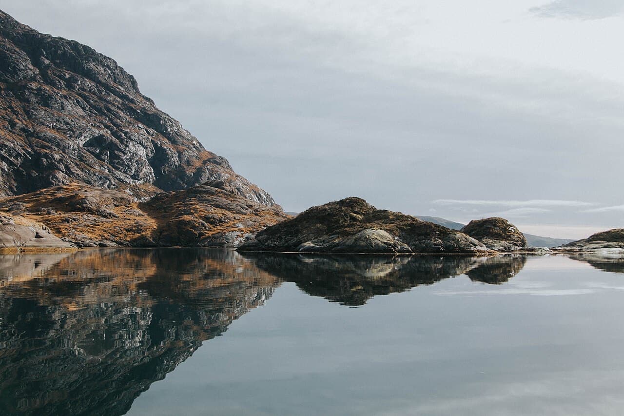 Rocky islets and the dark lower slopes of the Cuillin reflected in the mirror-still surface of Loch Coruisk under a grey sky.