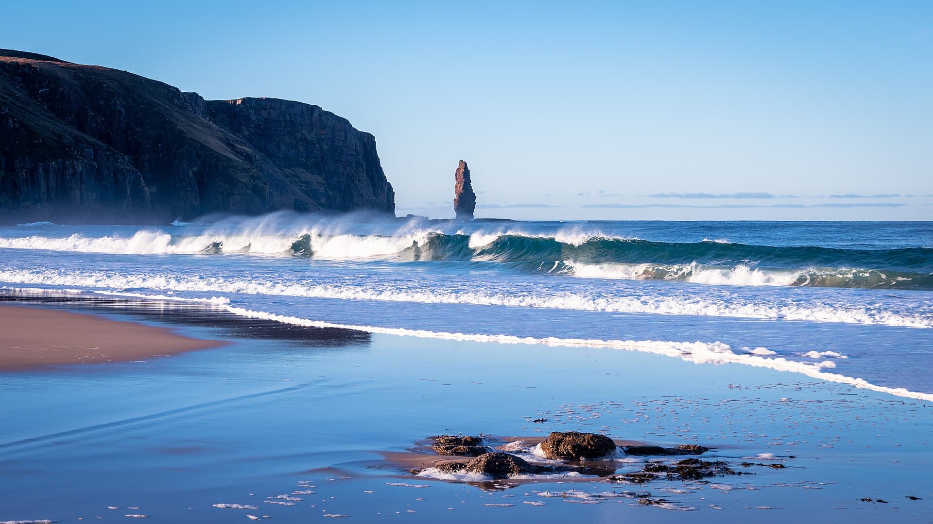 Teal breakers trail wind-blown spray across Sandwood Bay while the sea stack Am Buachaille rises between shadowed cliffs and the horizon, wet sand mirroring the sky.