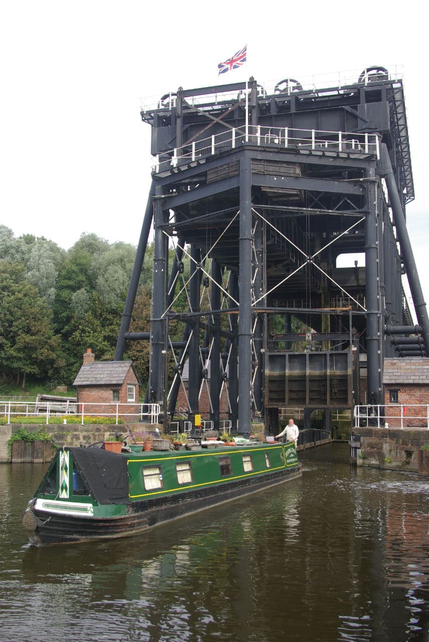A green narrowboat glides out of the Anderton Boat Lift's caisson onto the River Weaver, dwarfed by the black ironwork above.
