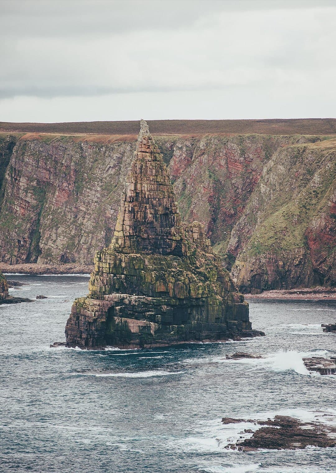 The great pyramid stack of Duncansby stands alone in the surf below striped sandstone cliffs.