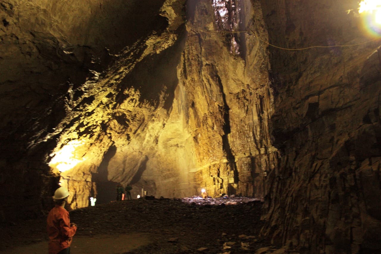 A caver in an orange oversuit and white helmet stands dwarfed at the edge of Gaping Gill's floodlit main chamber, tiny figures crossing the boulder floor far beyond.