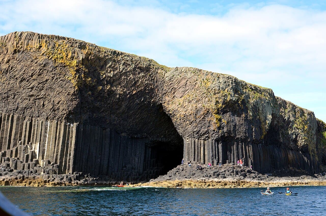 The arched mouth of Fingal's Cave opens in Staffa's columned basalt cliff, with sea kayakers paddling the water below.