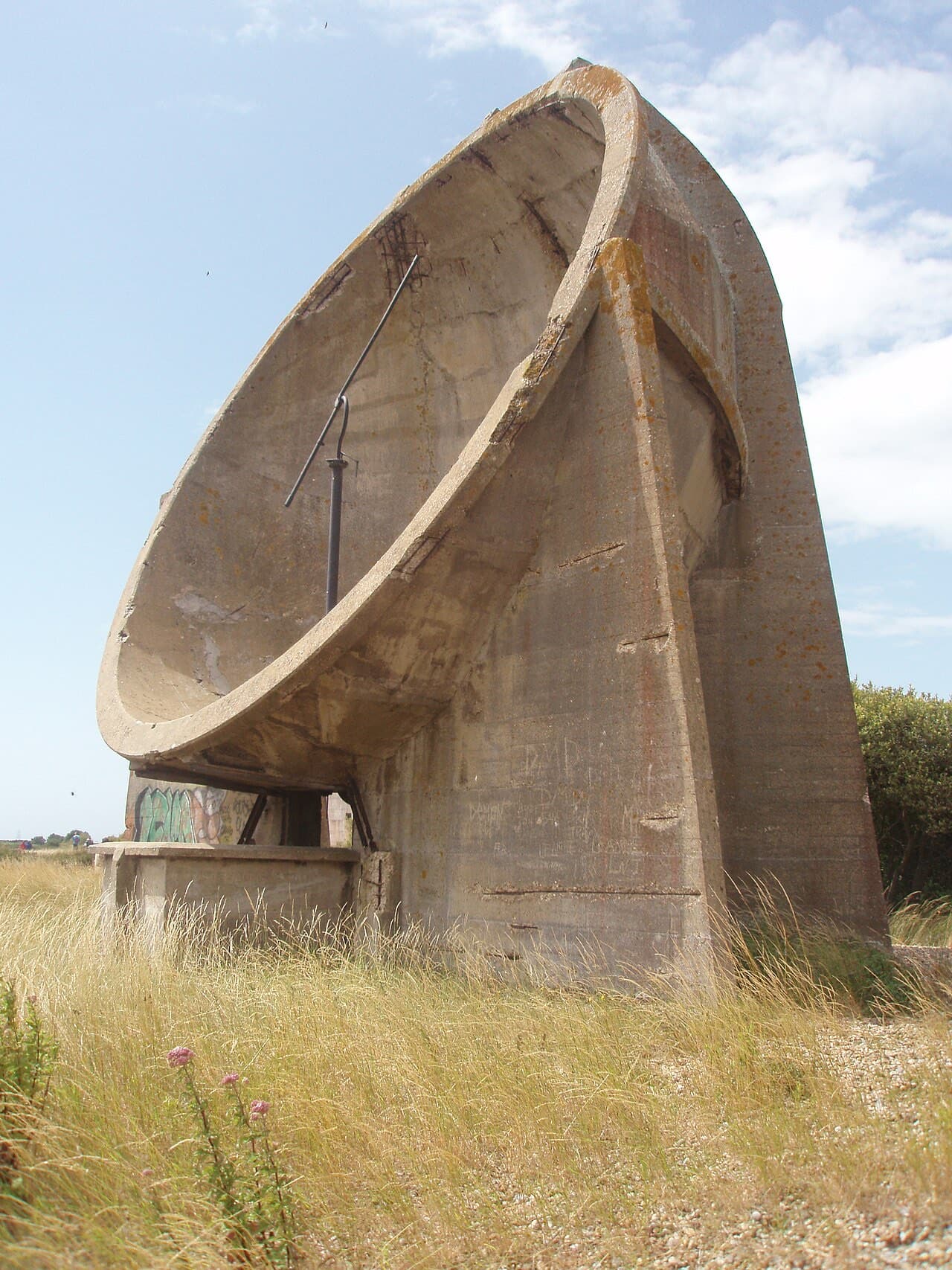 The thirty-foot concrete bowl of a Denge sound mirror tilts skyward above dry grass, its listening pole still in place.