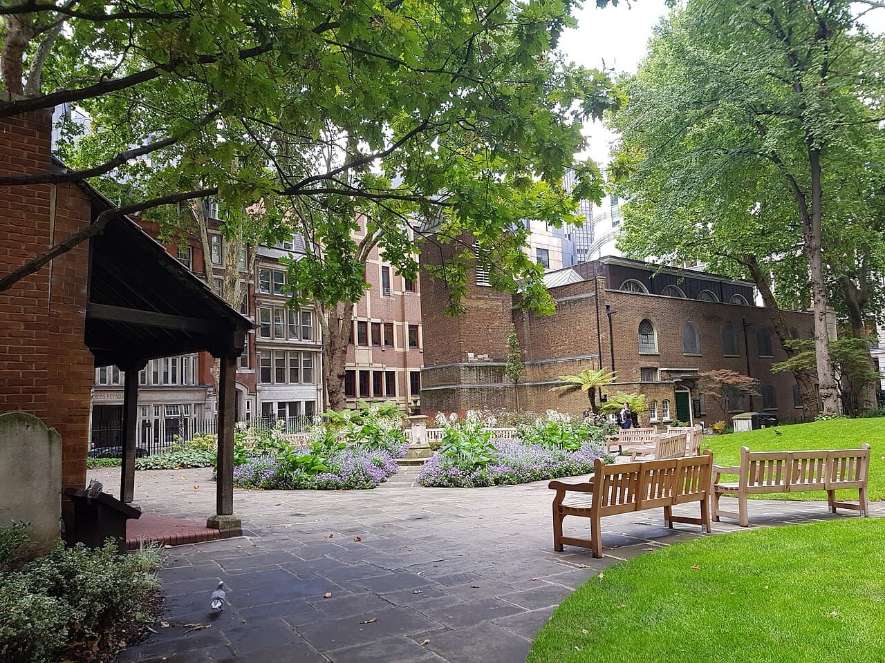 Leafy central garden of Postman's Park under an overhanging canopy, with wooden benches, beds of lavender-blue flowers around a small fountain, and the brick tower of St Botolph's Aldersgate beyond.