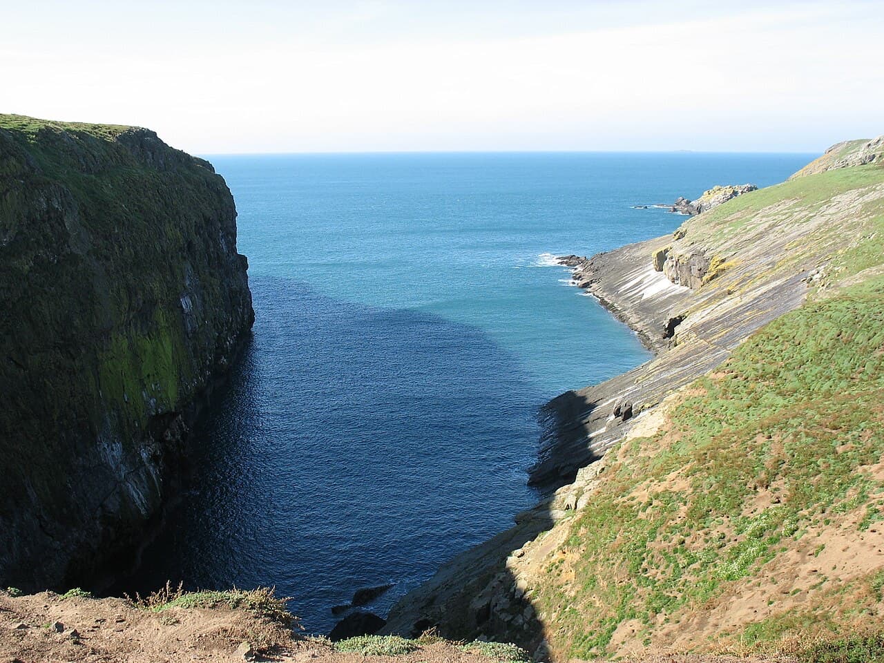 The sheer black cliff of the Wick on Skomer plunges into deep blue water beside sloping ledges of bare rock.