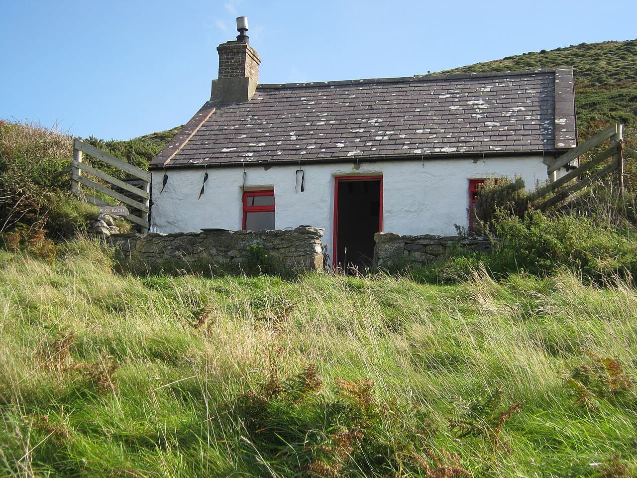 A whitewashed stone cottage with red-painted door and window frames stands in long grass below a hillside on Bardsey Island.