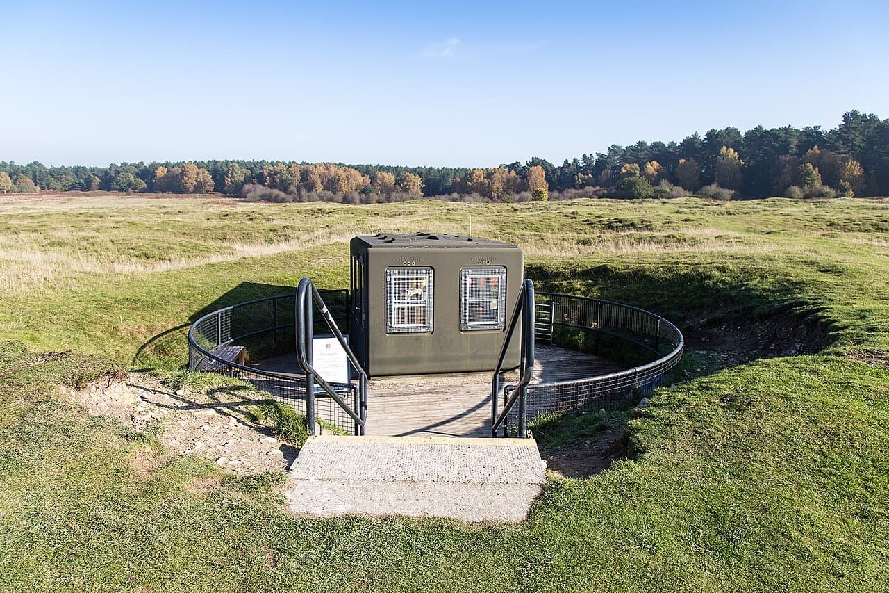 A small shelter and railed platform cap the open mineshaft at Grime's Graves, set among grassy craters with woodland behind.