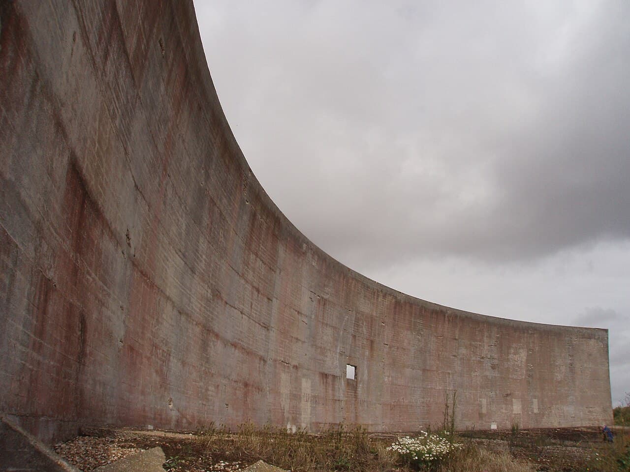 The curved two-hundred-foot wall mirror at Denge recedes under heavy grey cloud, its concrete stained and streaked with age.