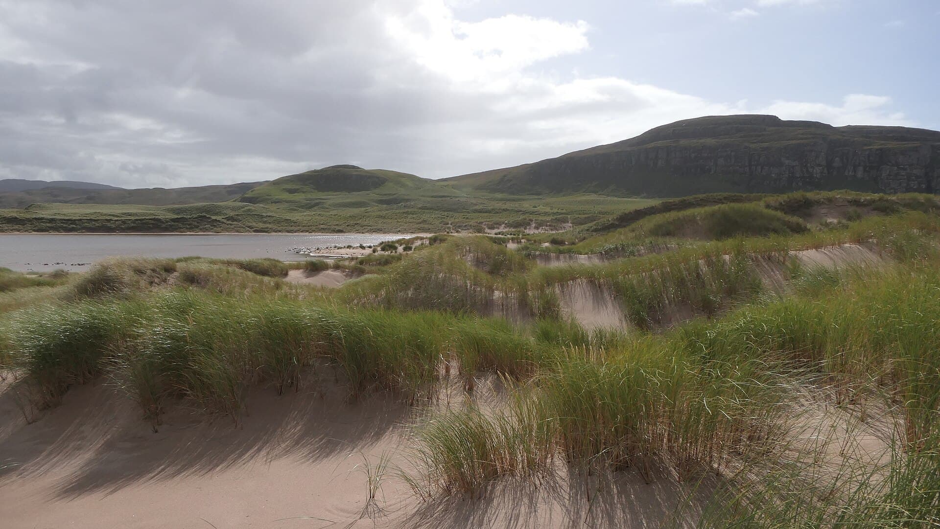 Wind-combed marram grass crests the dunes behind Sandwood Bay, with Sandwood Loch glinting silver beneath dark crags beyond.